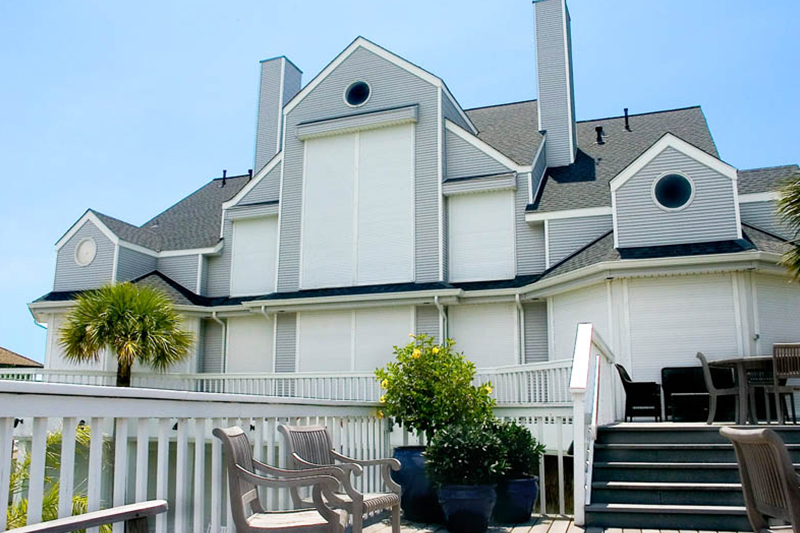 A two-story gray house with white trim and closed hurricane shutters, featuring a wooden deck, and outdoor furniture.