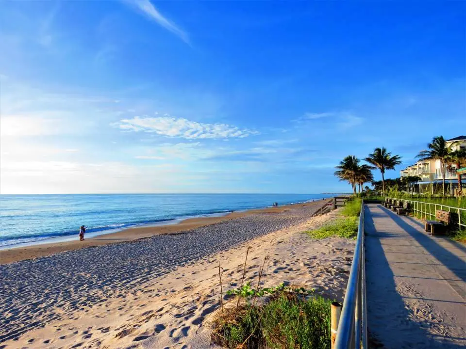 Sunlit beach scene with a sandy shore, calm blue ocean, and a boardwalk with palm trees on the right.