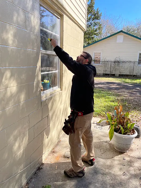 Man in a black jacket using a tool to work on a window on a beige exterior wall, with a tool belt and outdoor plants nearby, performing hurricane insurance discount brevard county