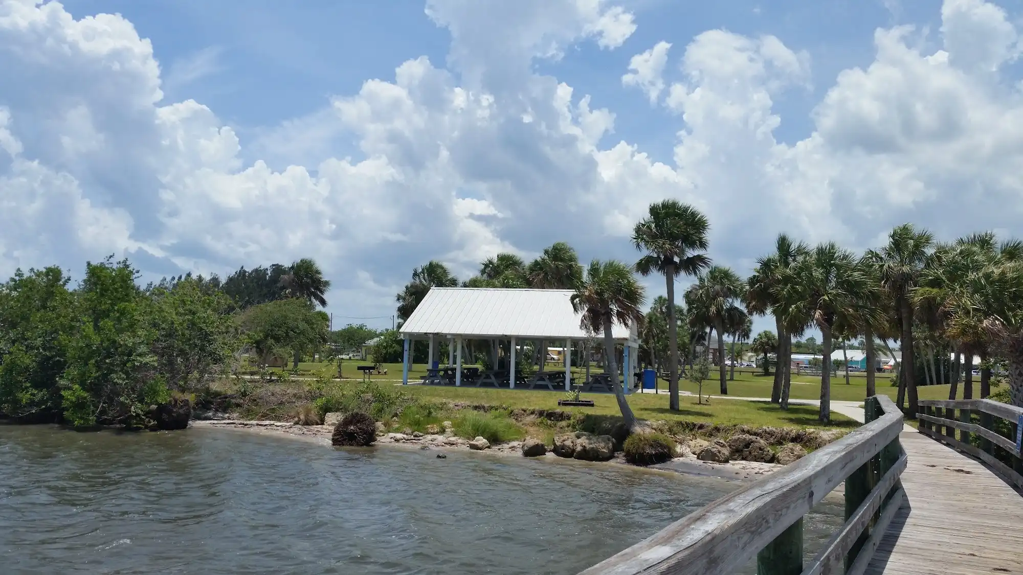 Waterfront park with a white-roofed pavilion, palm trees, and a wooden boardwalk along the shoreline under a cloudy sky.