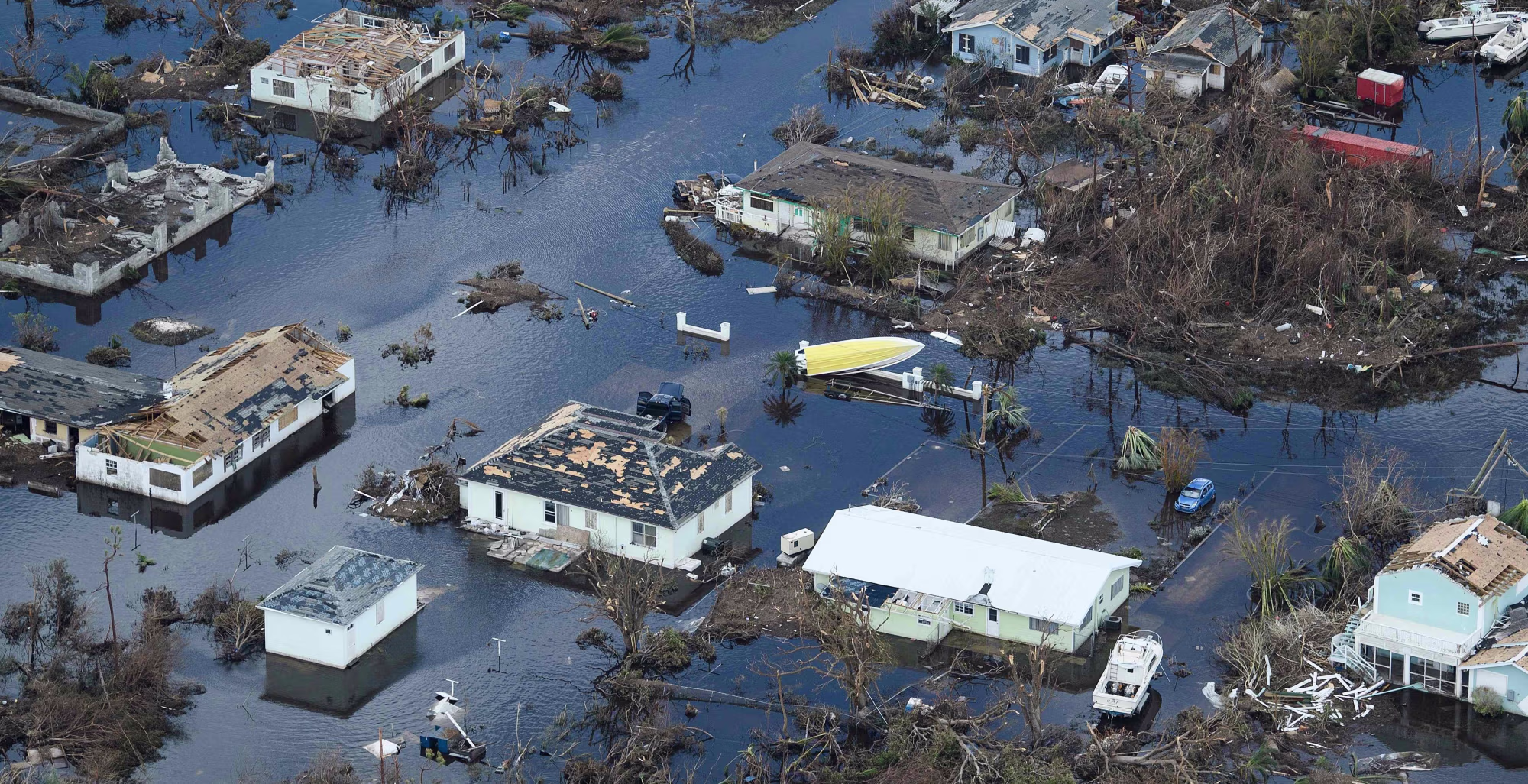 Aerial view of a flooded residential neighborhood with submerged houses, scattered debris, and fallen trees across the waterlogged streets.